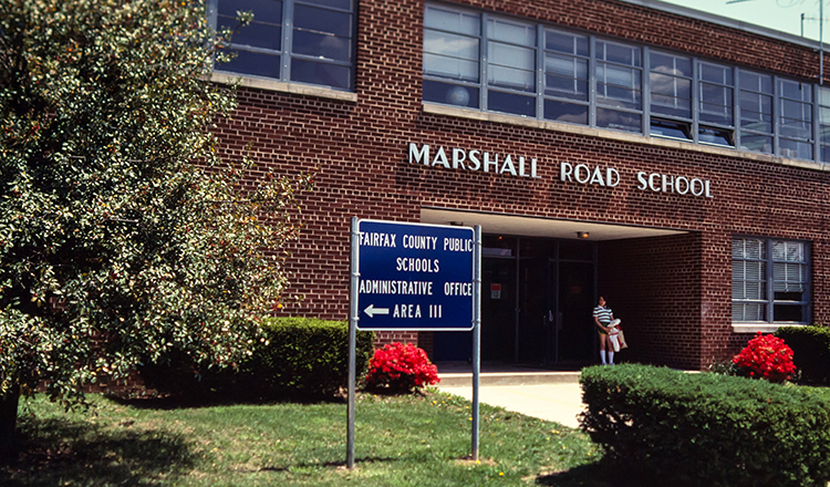 Color photograph from a 35 millimeter slide of the main entrance of Marshall Road Elementary School. The photograph is undated, but there is a student standing in the main entryway and because of the style of her clothing (blue and white striped shirt, shorts, and knee-high socks) it appears the picture was likely taken in the late 1970s or early 1980s. It is springtime and the flowers in front of the school are in bloom.  