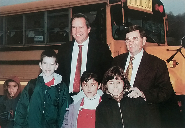Photograph of Marshall Road Center's principal John Marston with Marshall Road Elementary's principal David Meadows. They are standing in front of a school bus with a group of four children. 