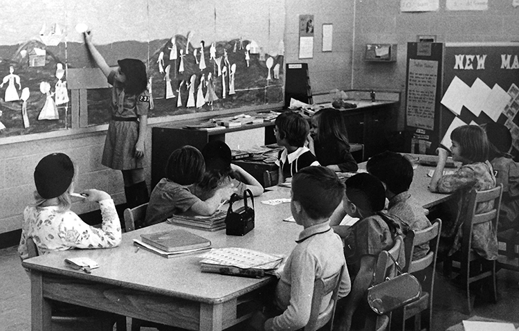 Black and white photograph of classroom 103, circa 1966. This was a primary-grade classroom, housing children in both first and second grades. A girl wearing a Brownie Scout uniform stands at a board along the wall which appears to have paper doll figures attached. A group of ten students, boys and girls, seated at a long wooden table look on. The children are seated on wooden chairs. 