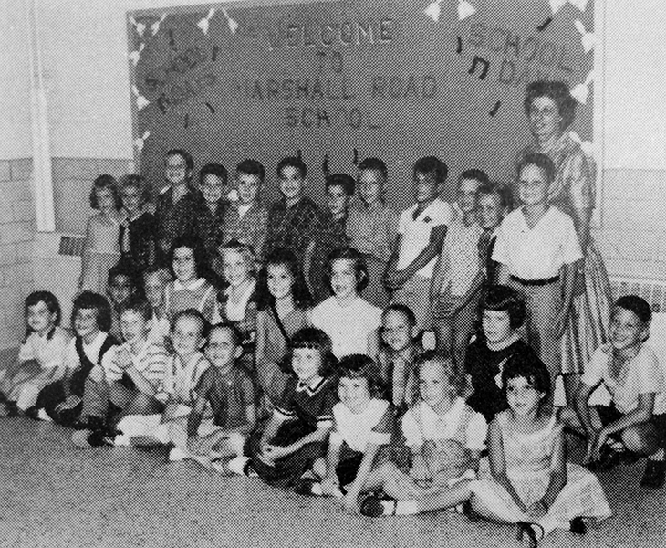 Black and white photograph of first grade teacher Mrs. Maude Foster with her students on the first day of school in 1961. There are thirty children pictured, boys and girls. They are photographed in a school hallway in front of a billboard that says Welcome to Marshall Road School. 